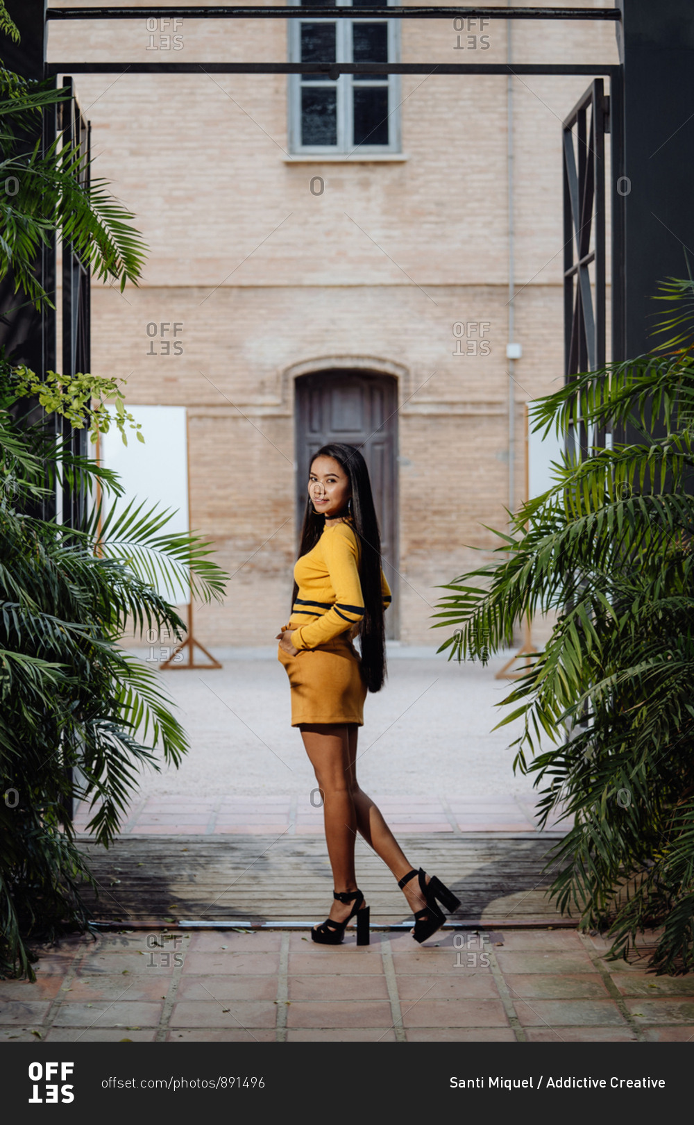 Side view of stylish trendy Asian woman with long dark hair in yellow short dress posing under decorative metal arch and looking at camera on background of building