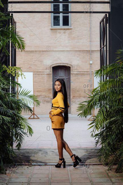 Side view of stylish trendy Asian woman with long dark hair in yellow short dress posing under decorative metal arch and looking at camera on background of building