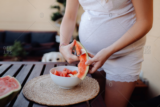 Crop pregnant female in white home clothes cutting watermelon to white bowl at terrace while dog sitting behind wooden table and watching
