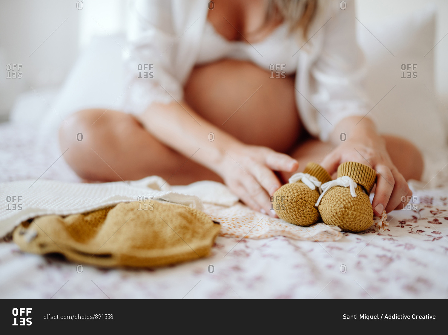 Crop faceless pregnant female dressed in white open blouse and bra showing clothes for unborn while sitting on bed with crossed legs