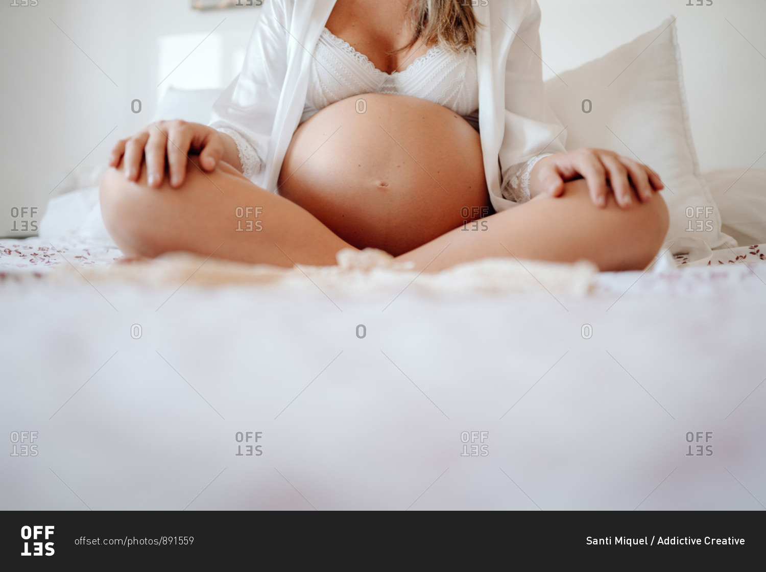 Crop faceless pregnant female dressed in white open blouse and bra sitting on bed with crossed legs