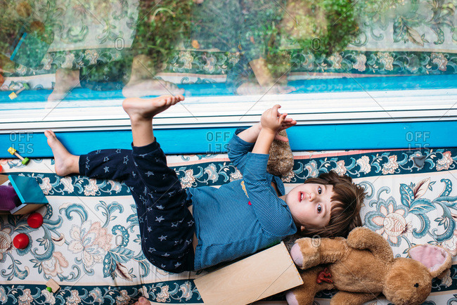 Little girl lying on a cushioned bench by a window with bare feet