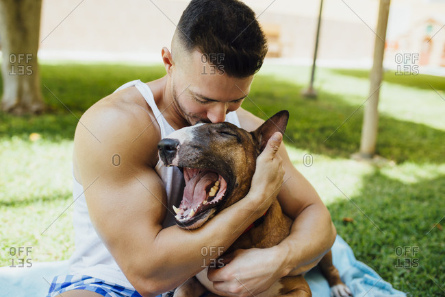 Muscular man sitting on blanket on a meadow hugging his dog