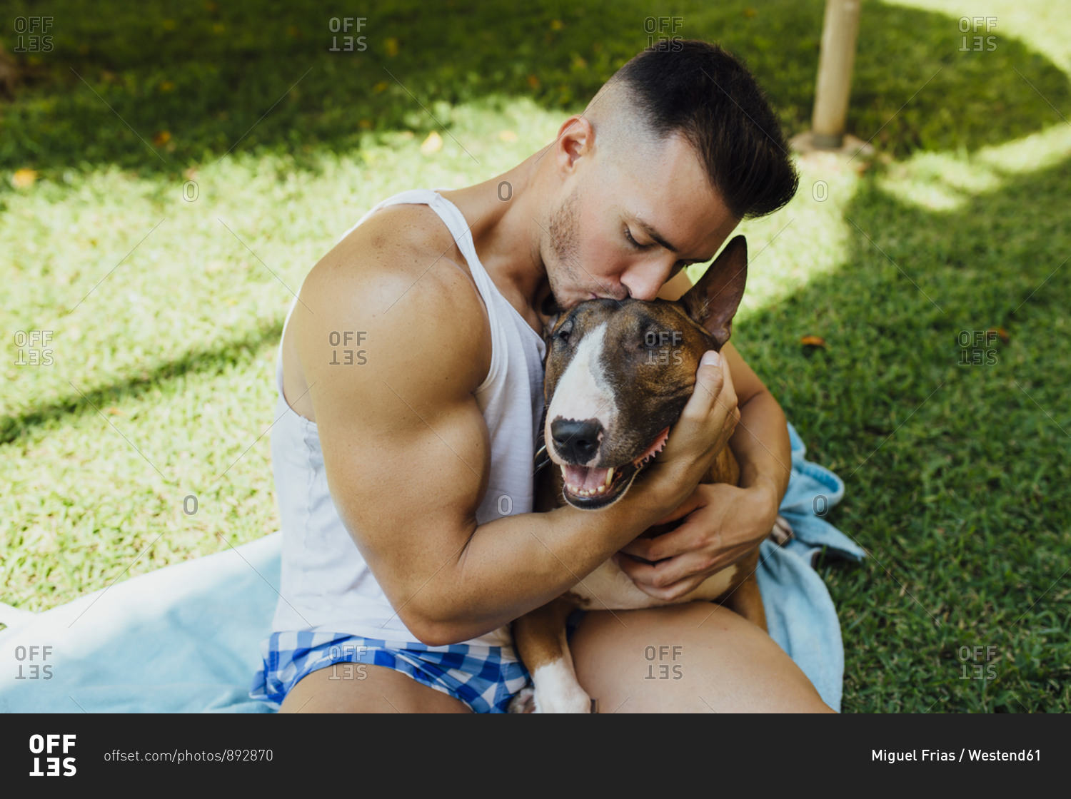Muscular man sitting on blanket on a meadow hugging his dog
