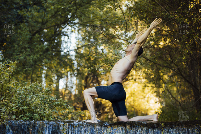 Young man practicing yoga on a waterfall- bending over back