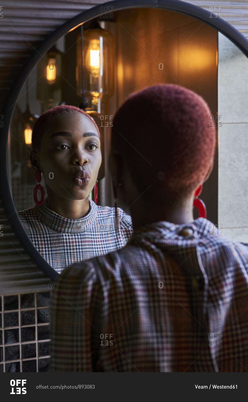 Young woman with short haircut looking to her reflection and sending a kiss in a round mirror
