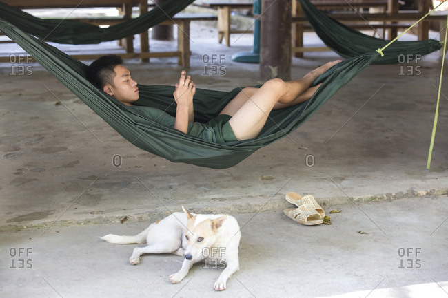 Vietnam, Ba Ria - Vung Tau - March 7, 2018: A young man lies in a hammock and looks at his phone. Dog beside him.