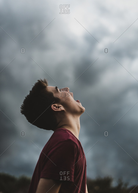 Teen boy looking up at dark cloudy sky with his mouth open wide.