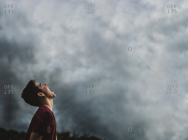 Teen boy looking up at dark cloudy sky with his mouth open wide.
