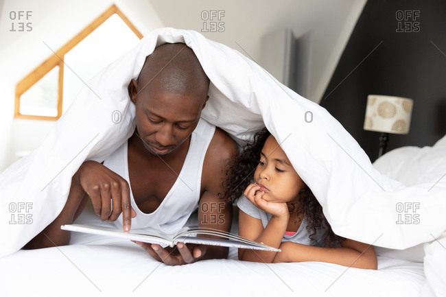 Front view of an African American man and his young daughter relaxing in the bedroom, lying under the covers and reading a book together