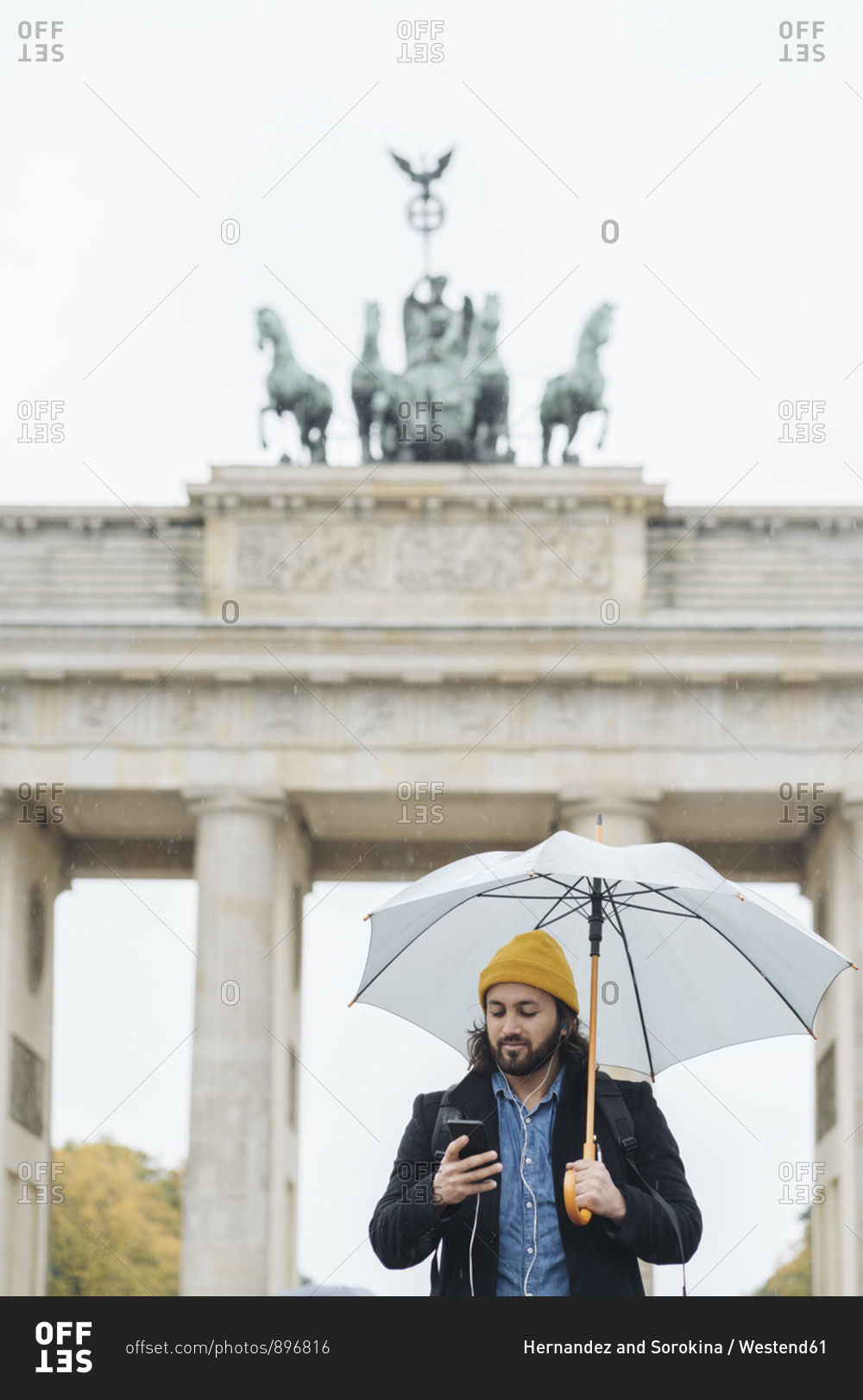 Man with umbrella using smartphone in front of Branderburg Gate- Berlin- Germany