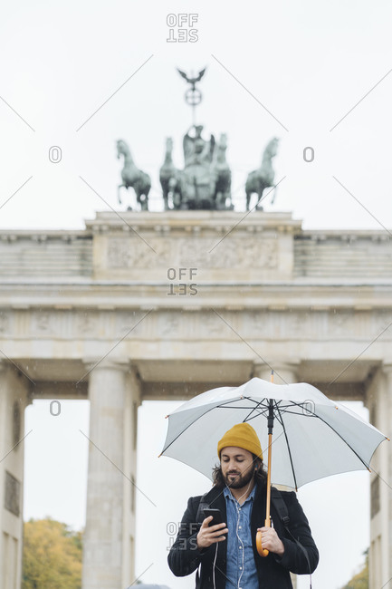 Man with umbrella using smartphone in front of Branderburg Gate- Berlin- Germany