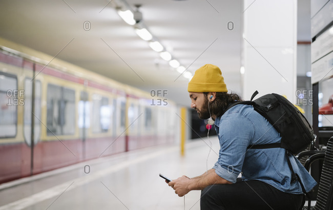 Man with backpack listening music with smartphone and earphones while waiting at platform- Berlin- Germany