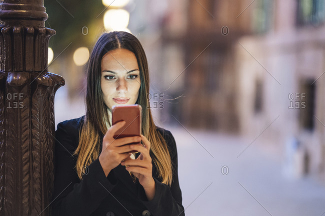Portrait of young woman leaning against lamp pole in the evening looking at cell phone