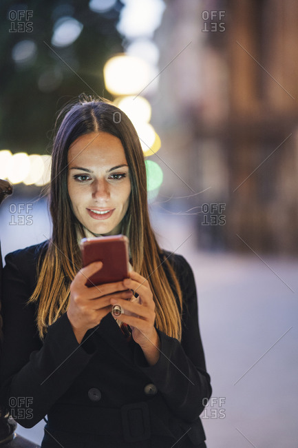 Portrait of smiling young woman leaning against lamp pole in the evening looking at cell phone