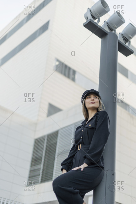 Young blond businesswoman wearing black sailor's cap and looking sideways- leaning on a lamp pole- low angle view