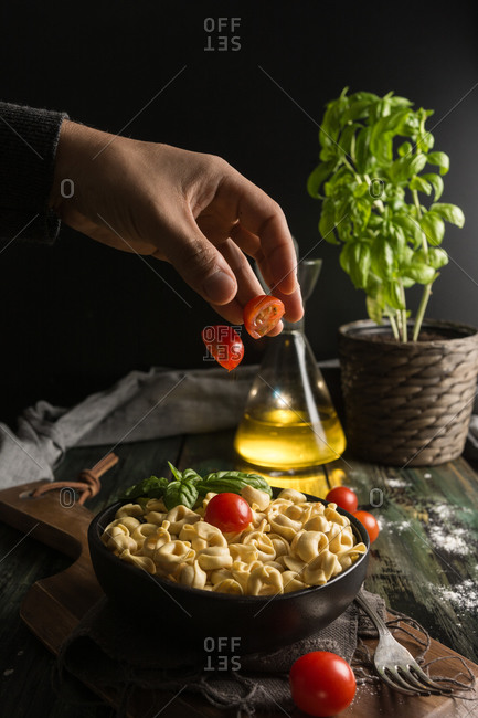Crop hand of man throwing cut fresh cherry tomato on black ceramic bowl with homemade ravioli placed on cutting board on table with glass jar with olive oil and basil plant