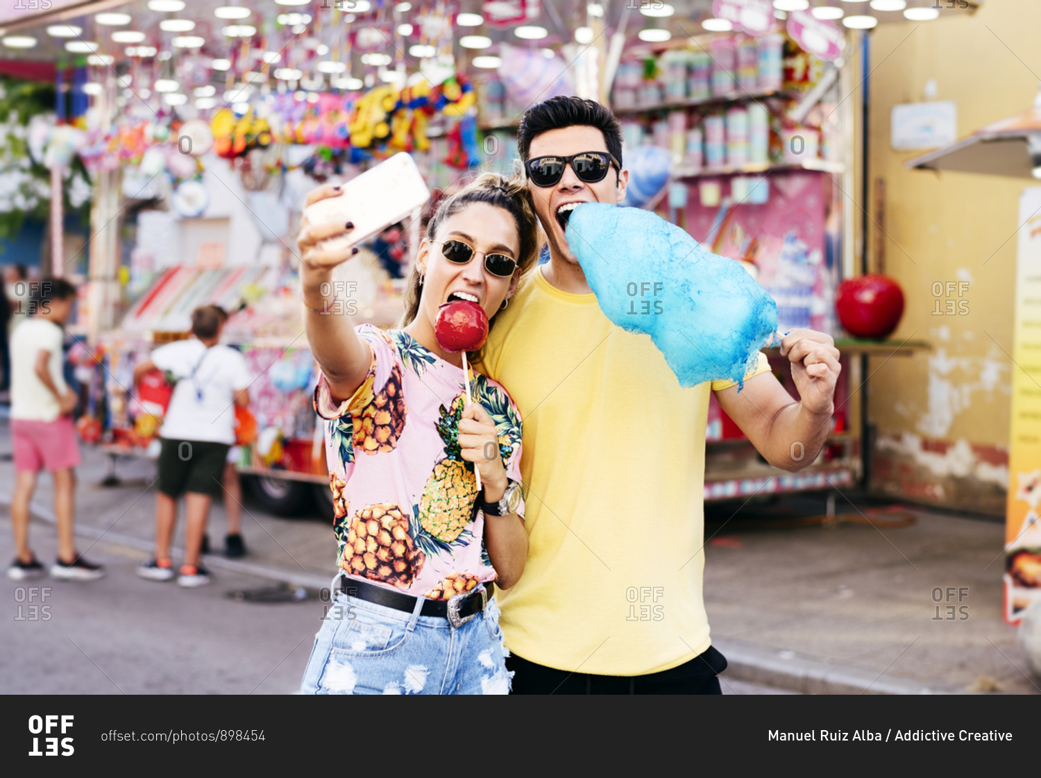 Joyful female in sunglasses and casual clothes using smartphone taking selfie with guy while eating candy apple and cotton candy on fair
