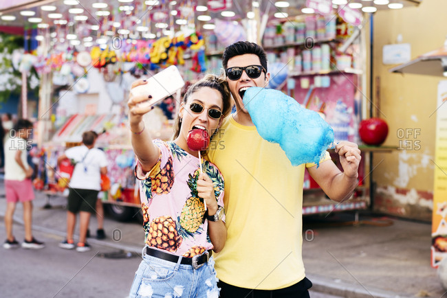 Joyful female in sunglasses and casual clothes using smartphone taking selfie with guy while eating candy apple and cotton candy on fair