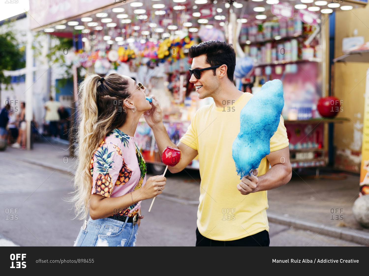 Happy male in sun glasses and casual clothes feeding young female with blue cotton candy while having fun on city fair