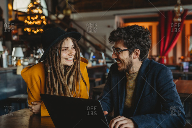 Smart man in glasses and stylish woman in hat looking at monitor while man typing on laptop in cafe