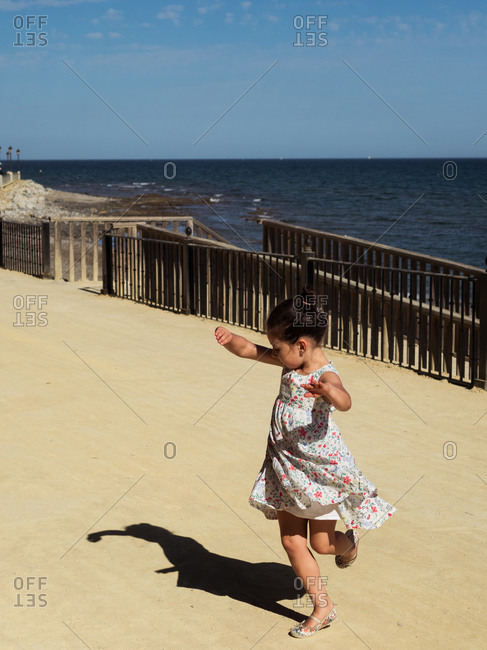 Full length little girl in floral dress dancing on embankment on sunny day near sea