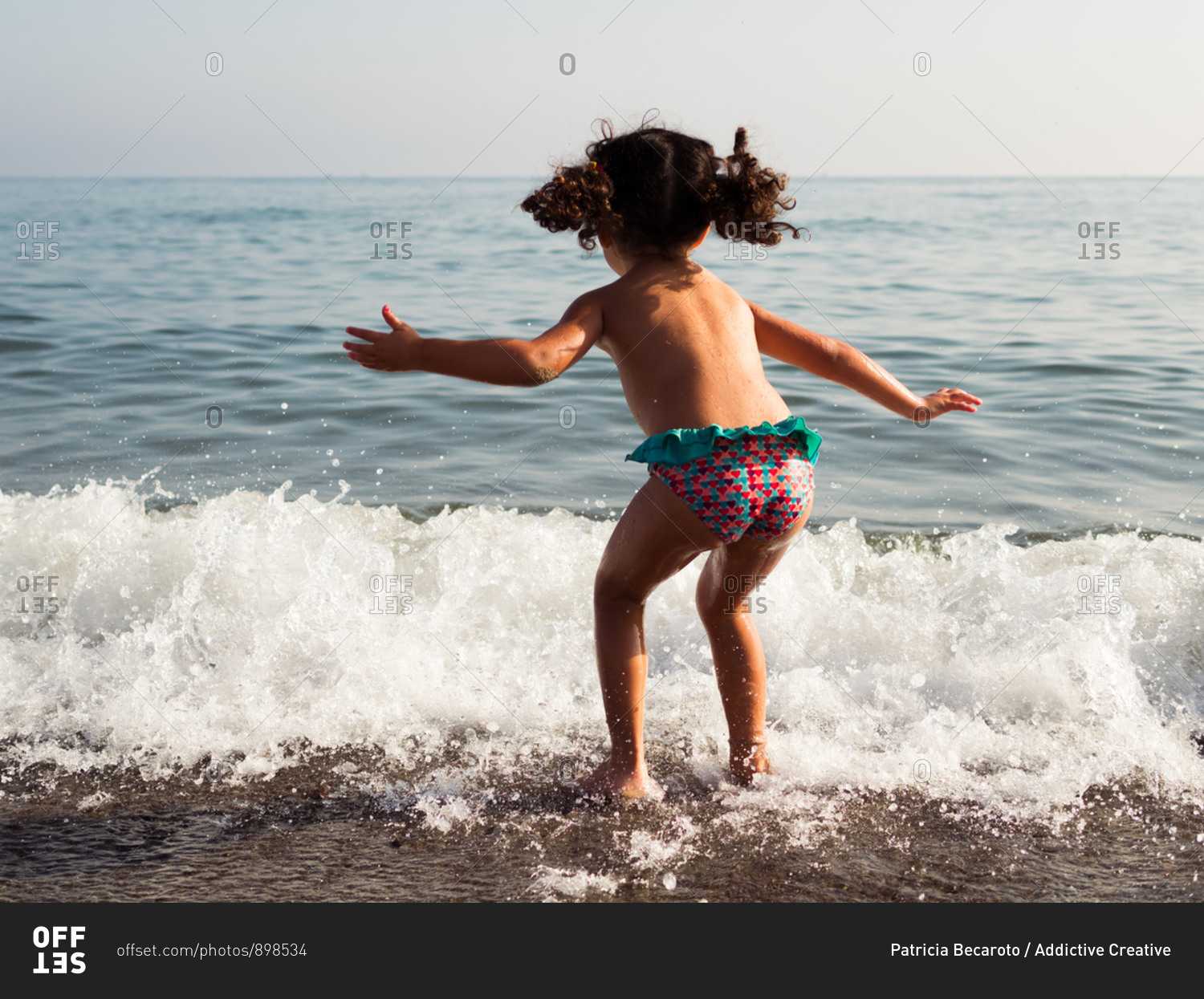 Back view of little girl in panties jumping into sea waves while having fun  on beach stock photo - OFFSET