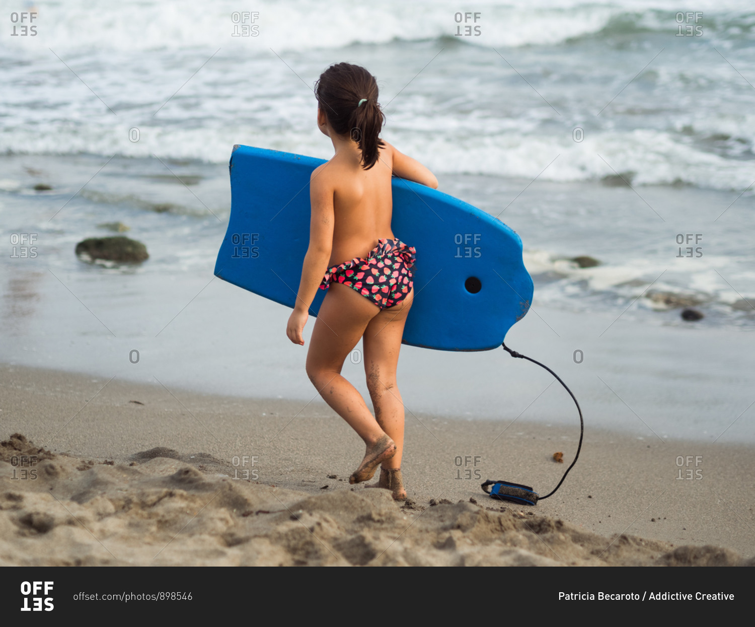 Back view of little girl in panties carrying blue swimming board while  walking on wet sand near sea stock photo - OFFSET