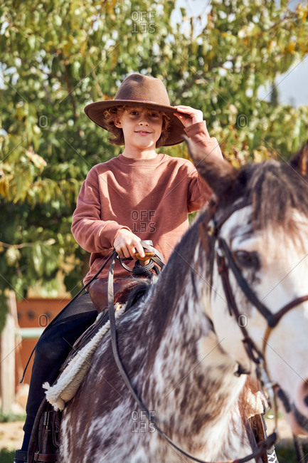 Happy little boy in hat looking at camera while riding his brown mottled horse