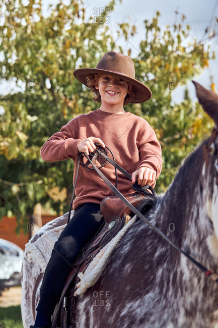 Happy little boy in hat looking at camera while riding his brown mottled horse