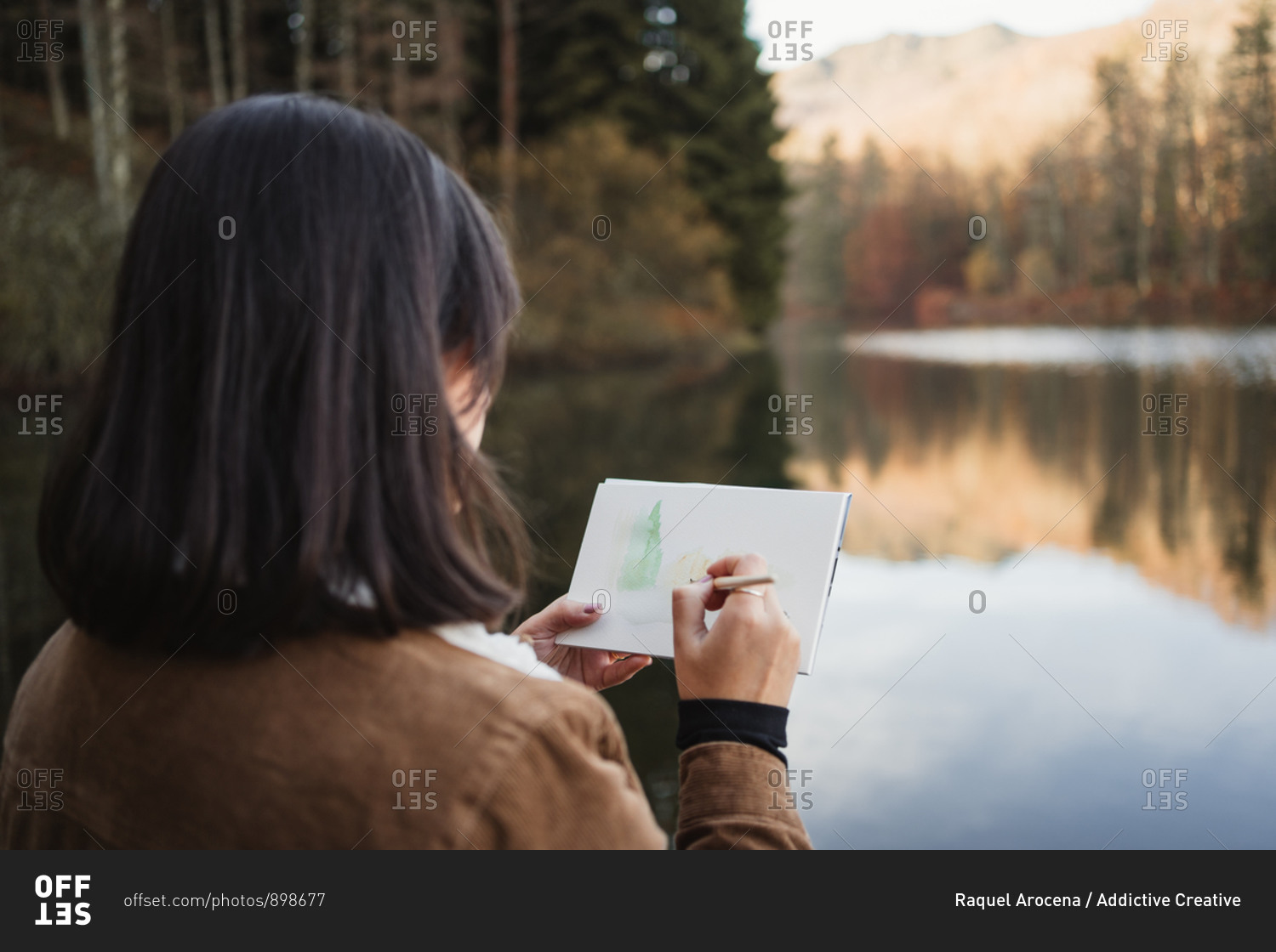 Back view of female artist standing next to beautiful lake surrounded with forest in autumn day and drawing in notebook