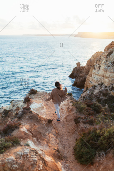 Back view of anonymous female in casual clothes walking towards edge of cliff near rippling sea in morning