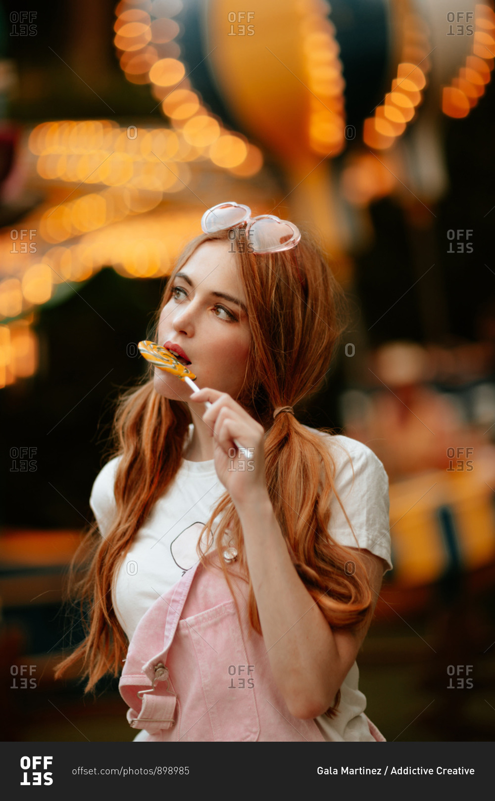 Charming red haired female teenager with ponytails and heart shaped eyeglasses on head standing in amusement park with illuminated attractions and eating lollipop