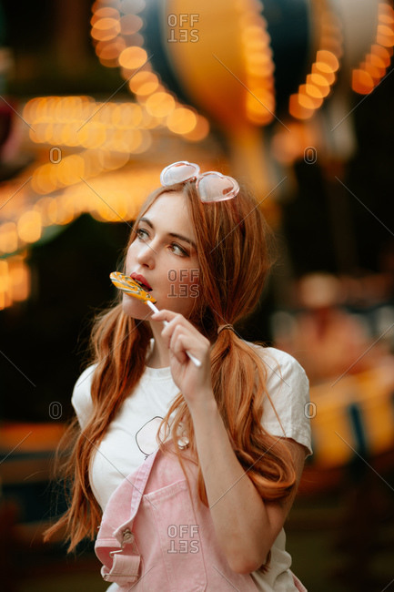 Charming red haired female teenager with ponytails and heart shaped eyeglasses on head standing in amusement park with illuminated attractions and eating lollipop