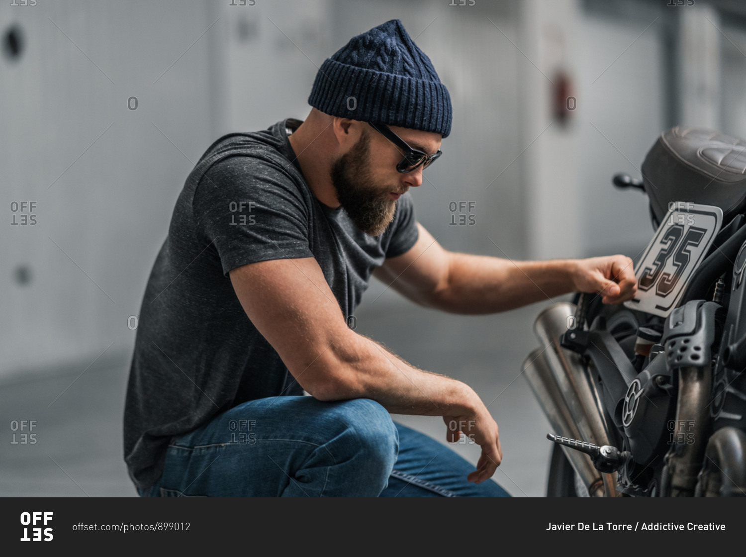 Bearded man in hat raising eyebrow and looking at camera while sitting near motorcycle on blurred background of contemporary garage