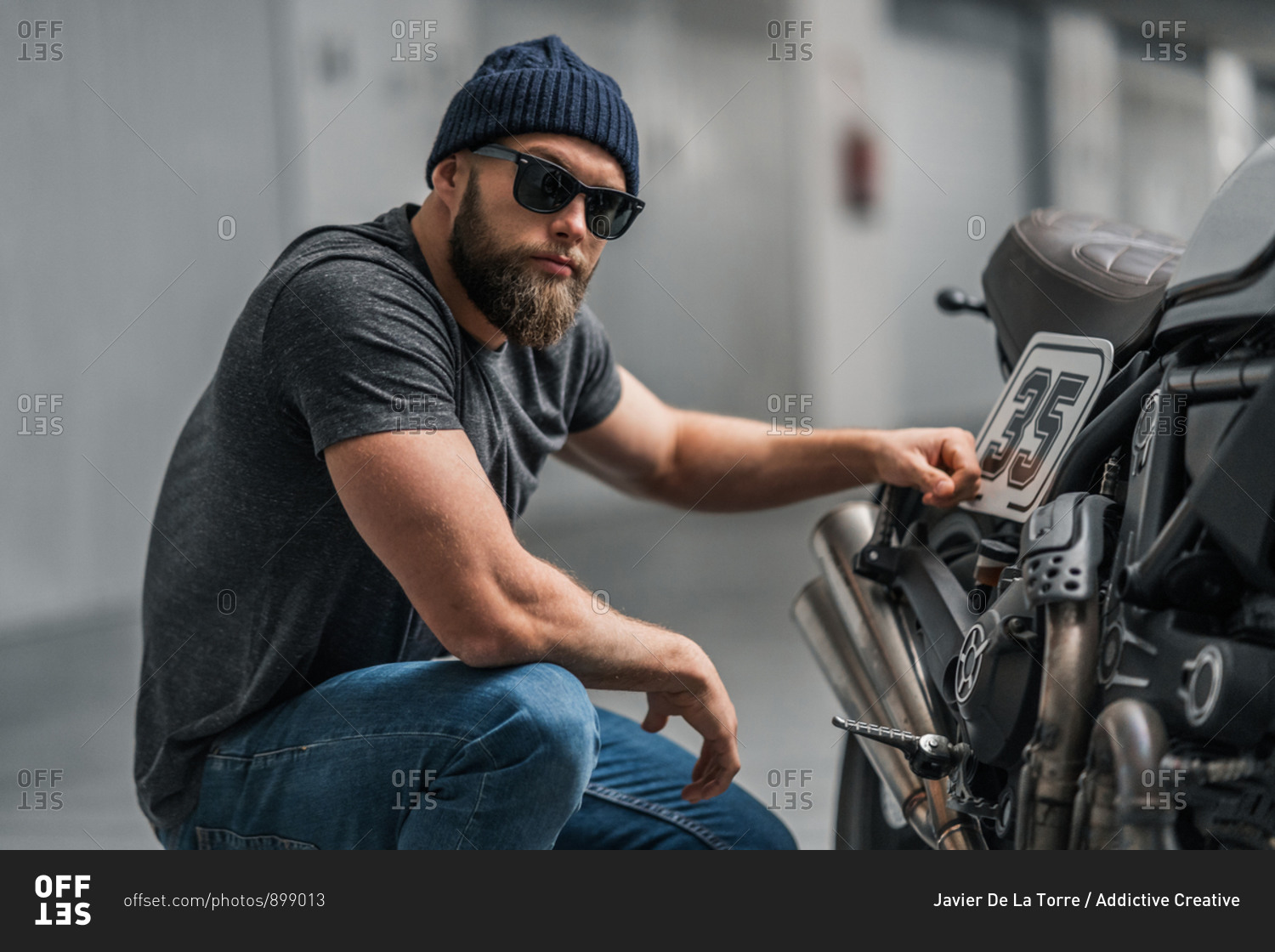 Bearded man in hat raising eyebrow and looking at camera while sitting near motorcycle on blurred background of contemporary garage
