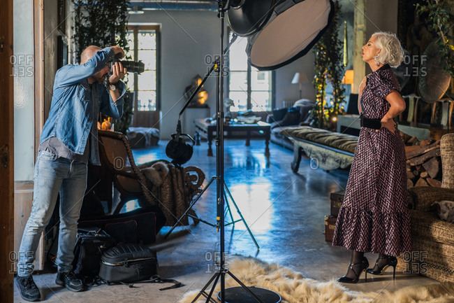 Low angle of professional photographer taking shot with camera of pensive gray haired lady in elegant dress standing against vintage rustic interior among off lighting equipment in photo studio