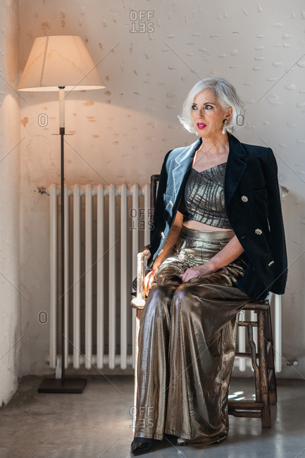 Low angle pensive lady in elegant black jacket and shiny stylish dress looking away and contemplating while sitting on wooden chair near white radiator and vintage standard lamp