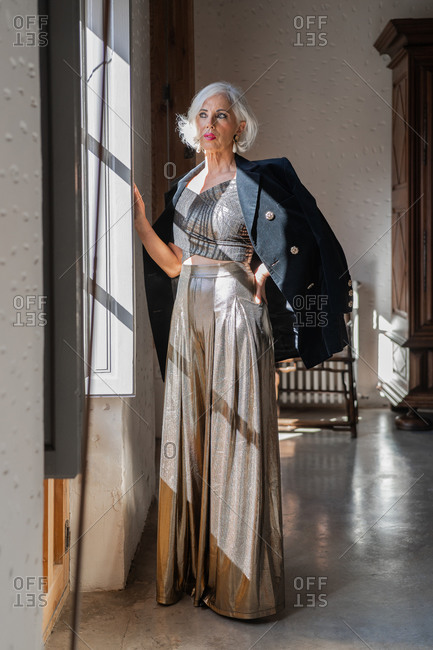 Low angle of pensive elderly female in stylish black jacket and golden shirt and trousers looking out opened window and contemplating while standing with hand on hip and leaning on frame against vintage interior