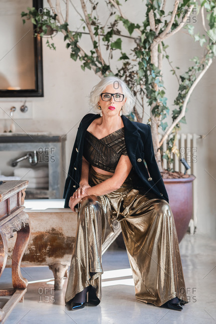 Calm senior confident lady in glasses and elegant clothes looking at camera while sitting on shabby bathtub edge among rich retro interior including old furniture and big green plotted tree in spacious room of country house