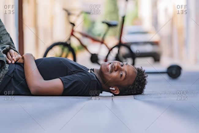 Side view of happy youthful African American man lying on asphalt road in street looking at camera