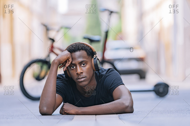 Concentrated youthful African American man wearing headphones listening to music while lying on asphalt road in street