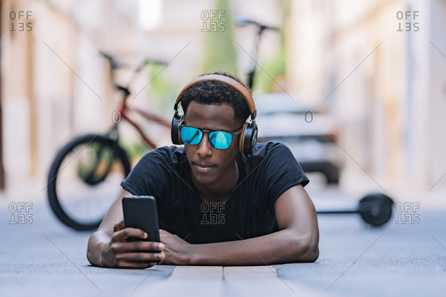 Concentrated youthful African American man in sunglasses wearing headphones and listening to music on the mobile phone while lying on asphalt road in street