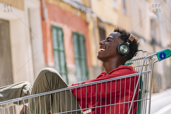 Side view of pensive youthful African American male teenager in casual clothes looking at camera while sitting in metal shopping trolley in street