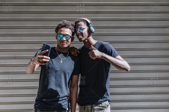 Cool youthful African American male teenagers in sunglasses taking picture with mobile phone while standing in sunlight in street