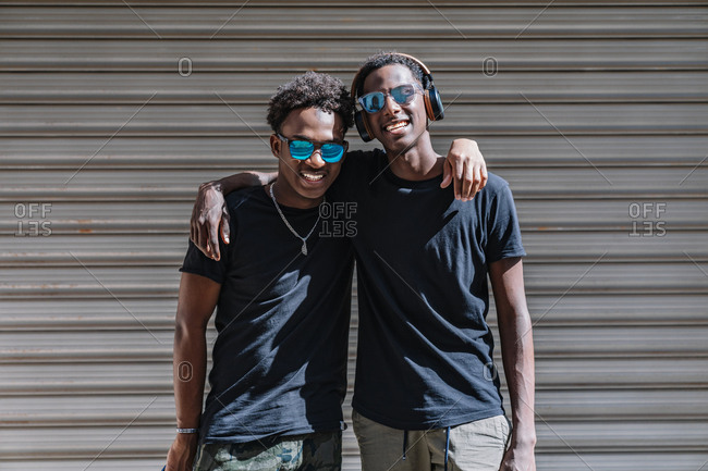Happy youthful African American male teenagers in sunglasses enjoying pastime together while standing in sunlight in street