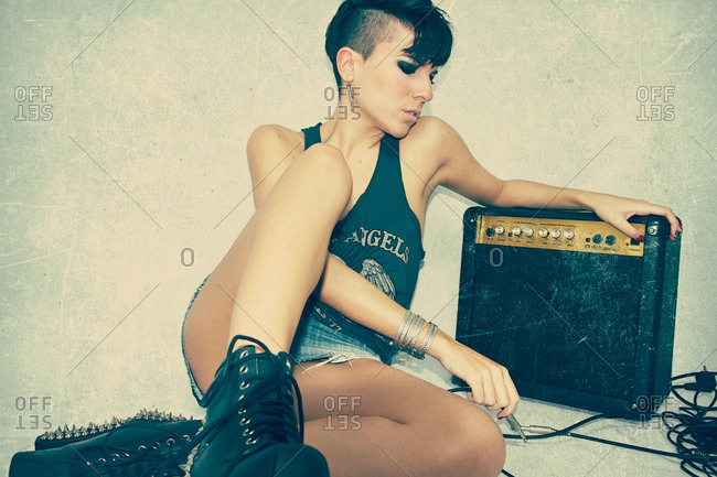 Thoughtful youthful woman in rocker clothes and with modern hairstyle sitting on floor with retro guitar combo amplifier in studio