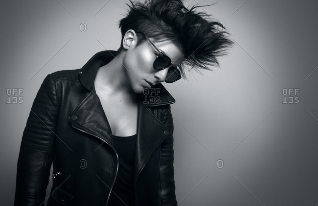 Independent serious youthful woman with short dark hair and sunglasses wearing biker jacket and looking down in studio on gray background