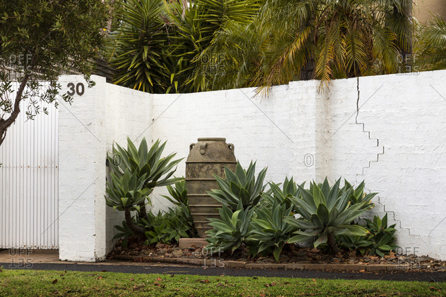 Balaclava, Victoria, Australia - June 22, 2019: Decorative large ancient clay amphora placed next to white stone wall with green tropical plants