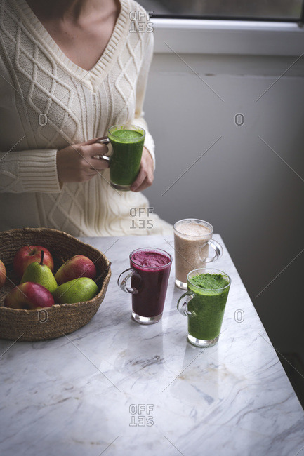 Woman sitting at the table holding a green smoothie in her hands and there are other smoothies in glasses on the table in front of her